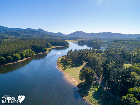 Aerial view of lake with boat, surrounded by trees and mountains on a sunny day.