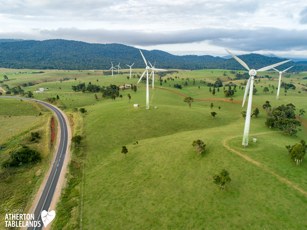 Wind turbines in a green field with a road; mountains in the background under a cloudy sky.