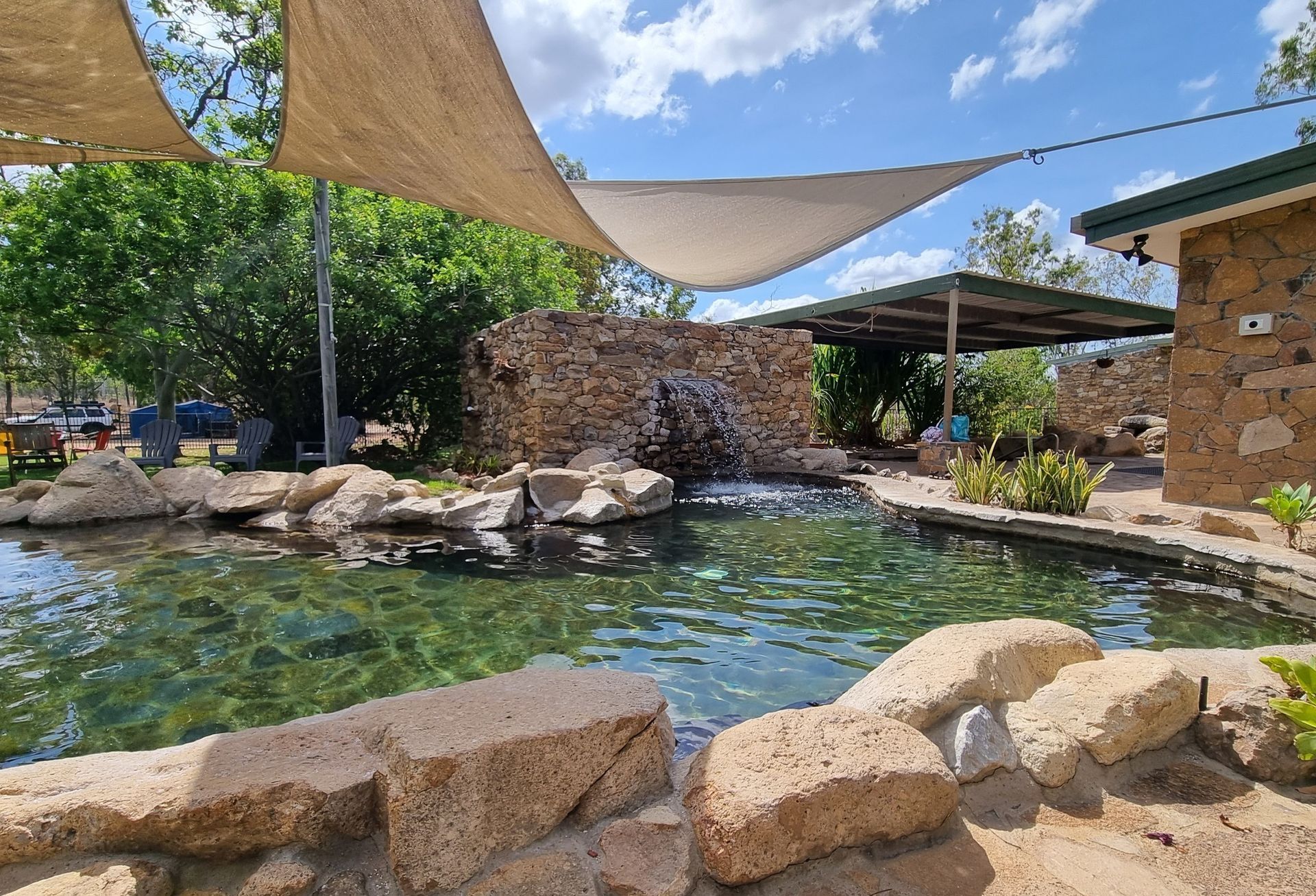 Pond with waterfall, rocks, and shaded seating area under a blue sky.