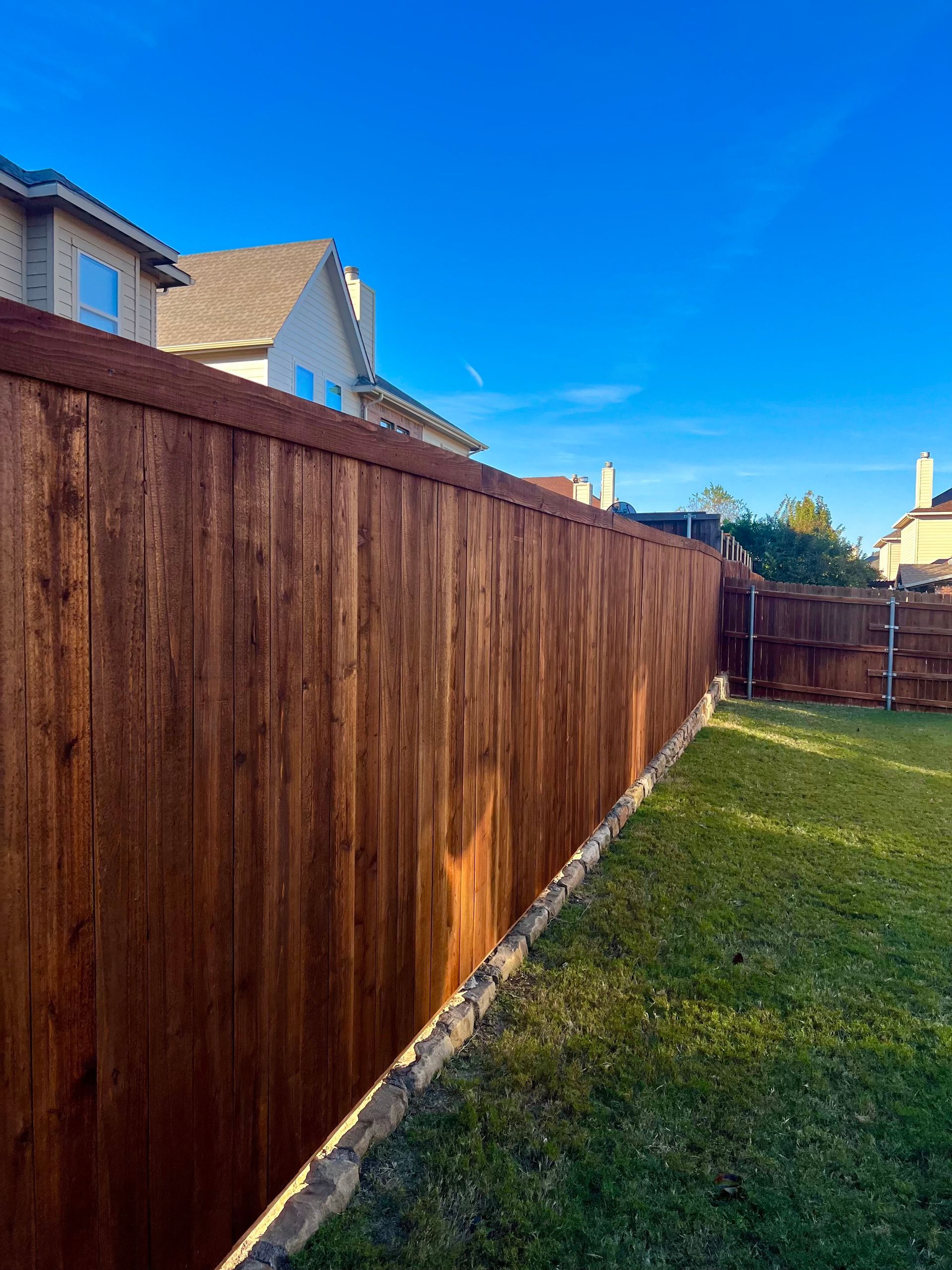 Wooden fence in a backyard with green grass and a house under a clear blue sky.