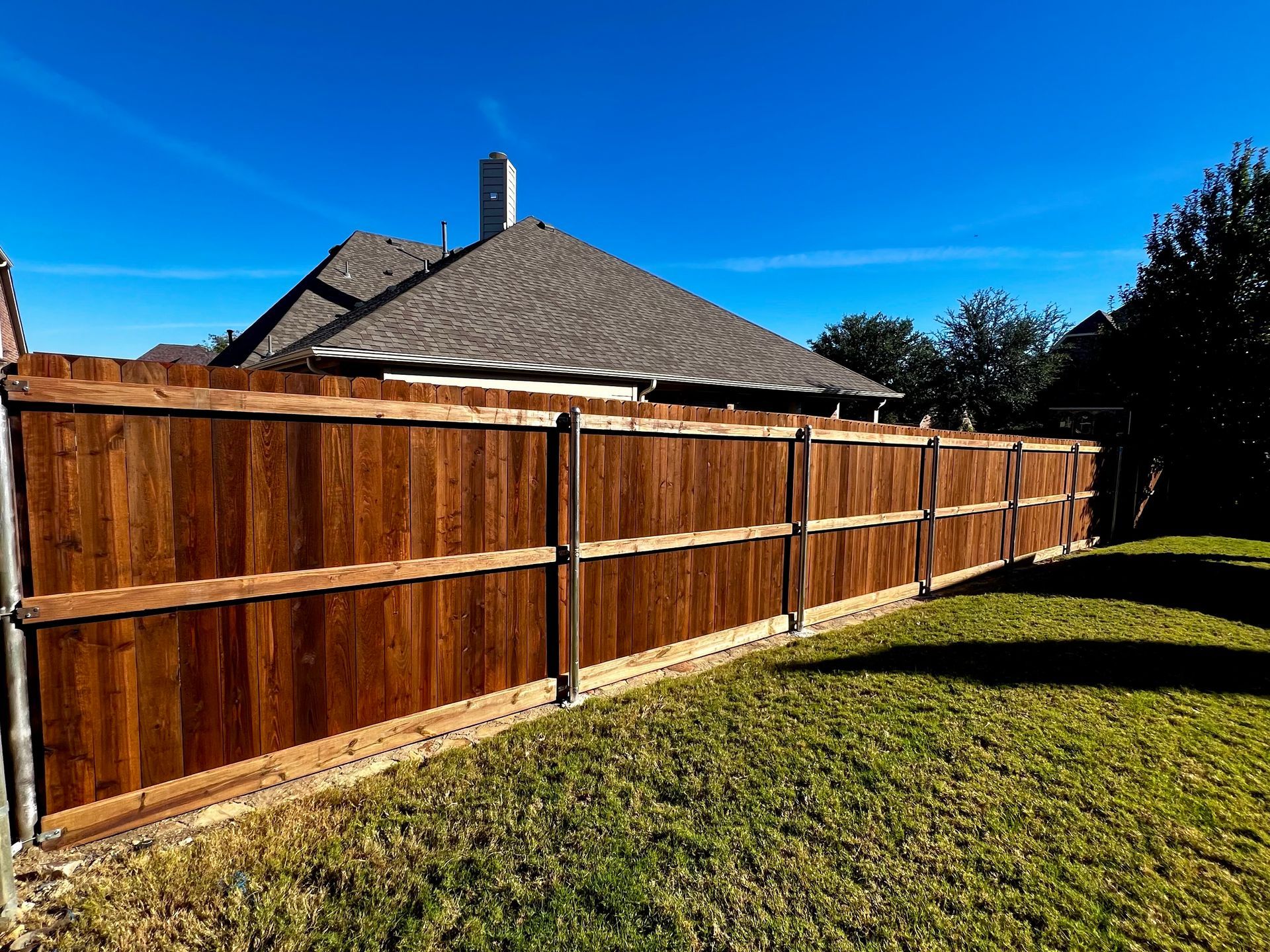 Wooden fence in backyard, brown stained with metal posts, green grass and blue sky.