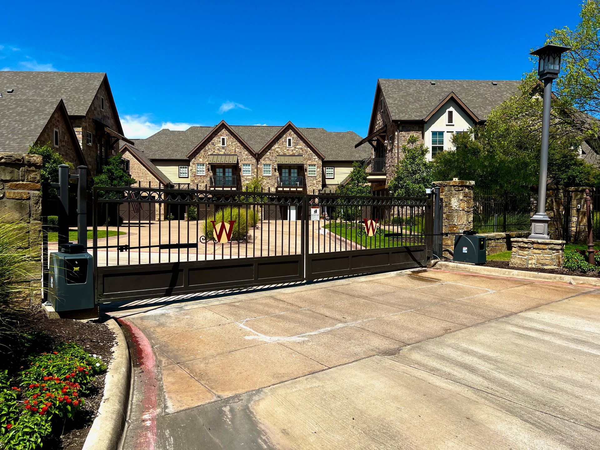 Gated community entrance with stone buildings, black gate, street lamp, blue sky.