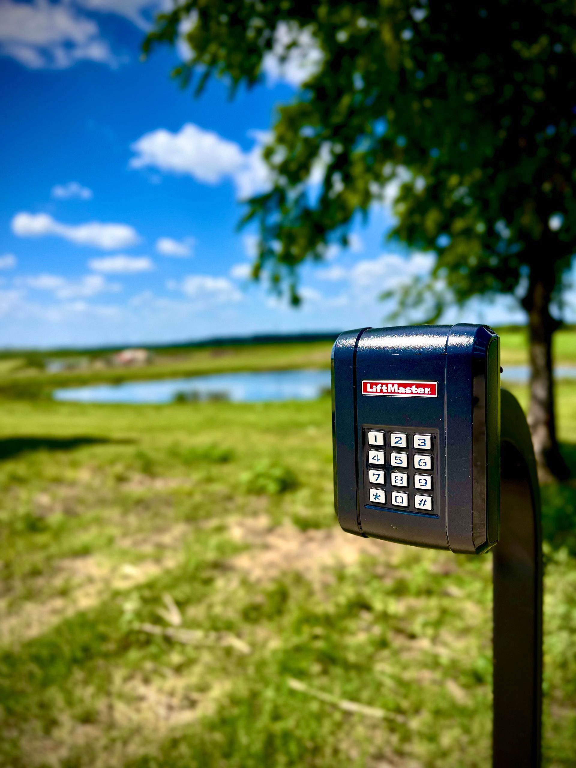 Black keypad on a post with a scenic background of green fields, a pond, and a blue sky.