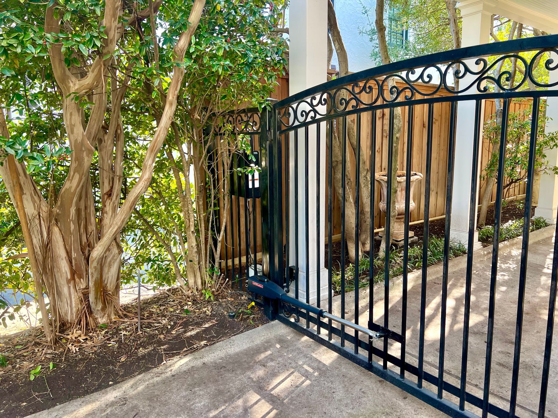 A wrought iron gate is open to a driveway with trees in the background.