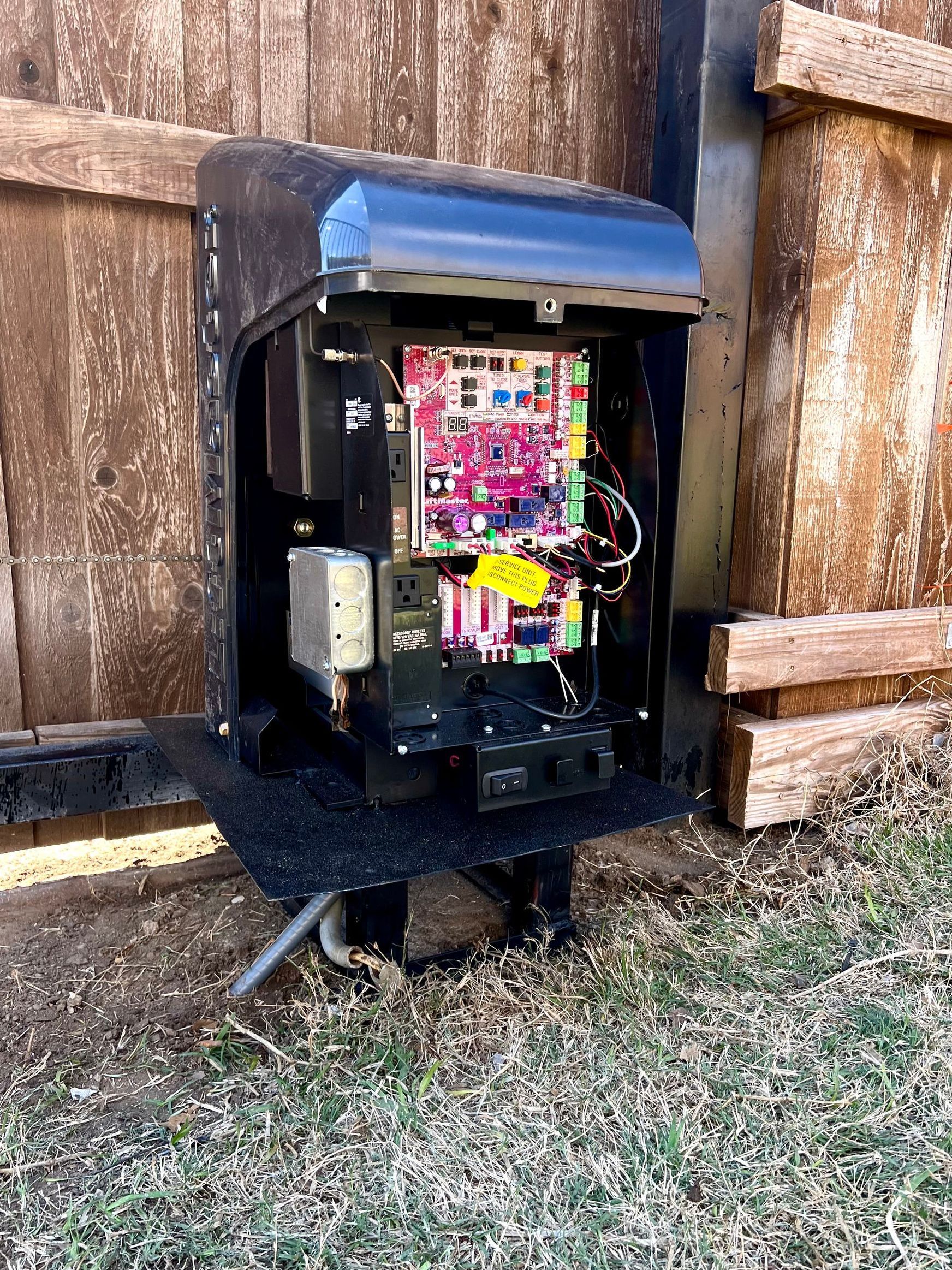 A electrical box is sitting in the grass next to a wooden fence.