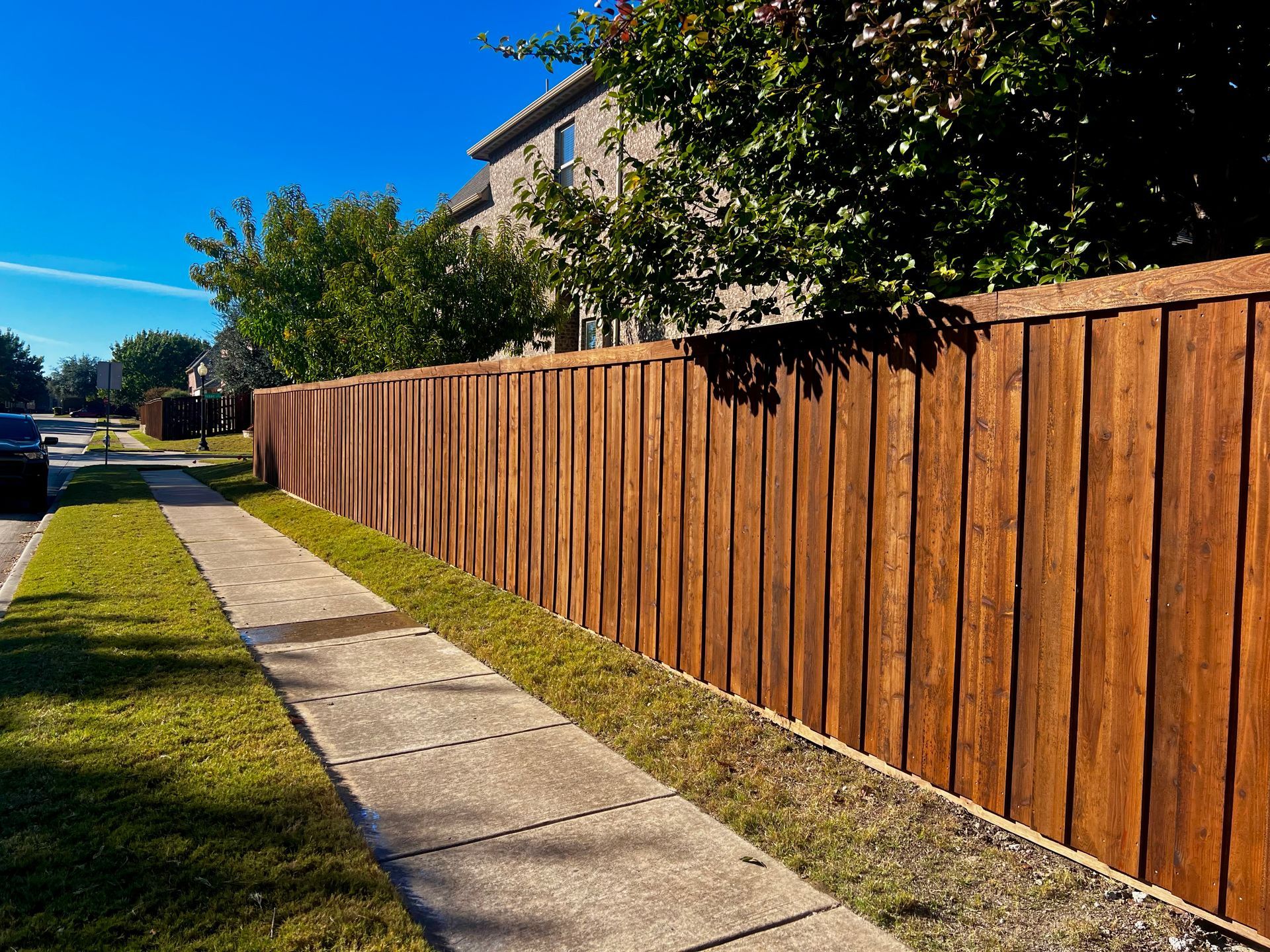 Brown wooden fence alongside a sidewalk and green grass, with a building and trees in the background under a blue sky.
