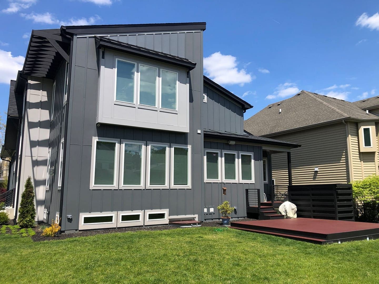 Modern two-story house with gray siding, large windows, and a red deck, in a sunny backyard with green grass.