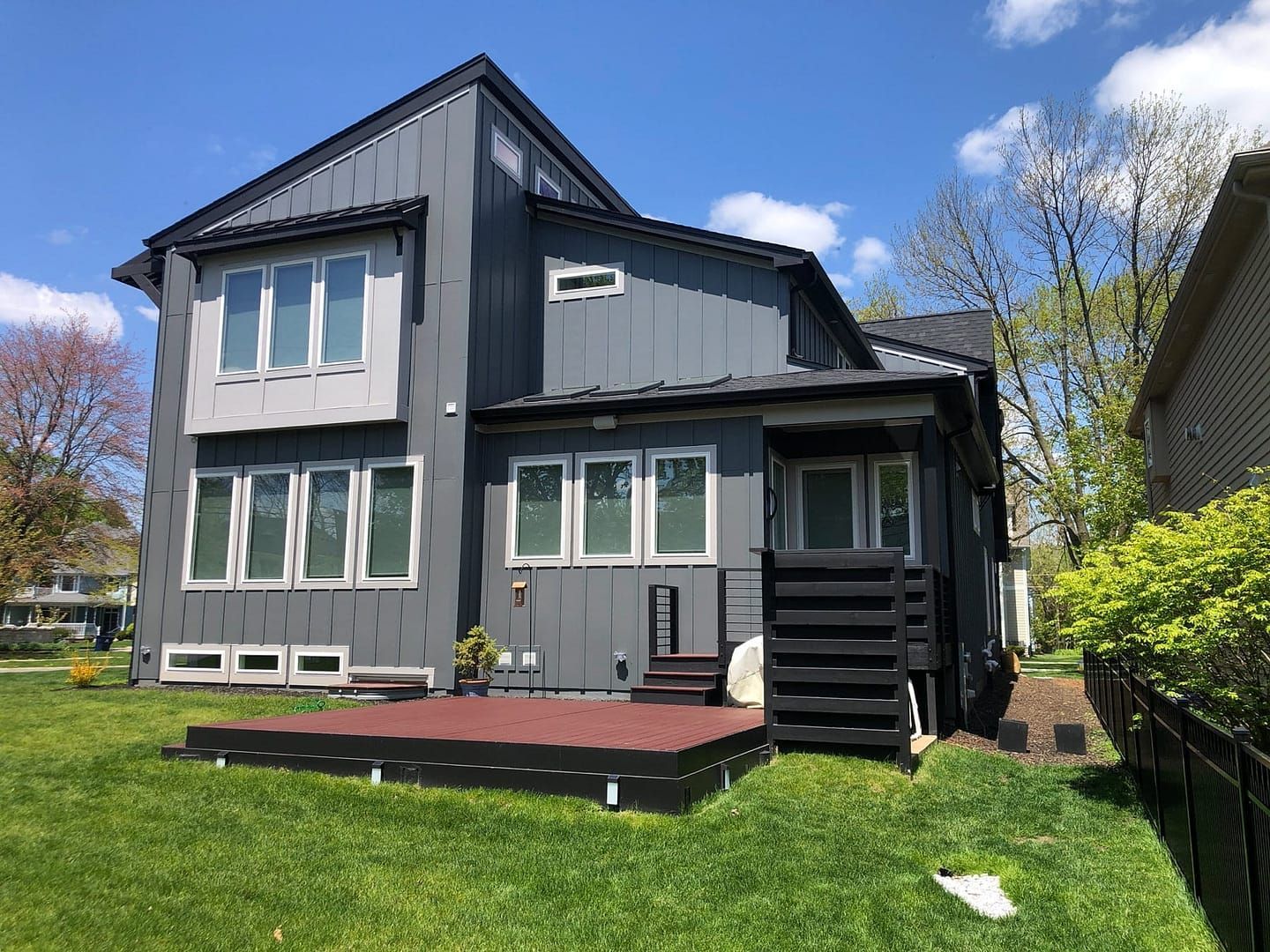 Modern gray house with black roof, deck, and stairs, viewed from a grassy yard.