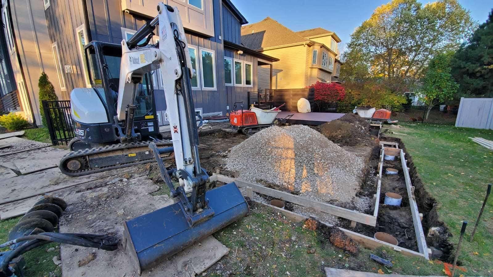 An excavator in a backyard, digging near a deck with gravel, soil, and wood forms; construction site.