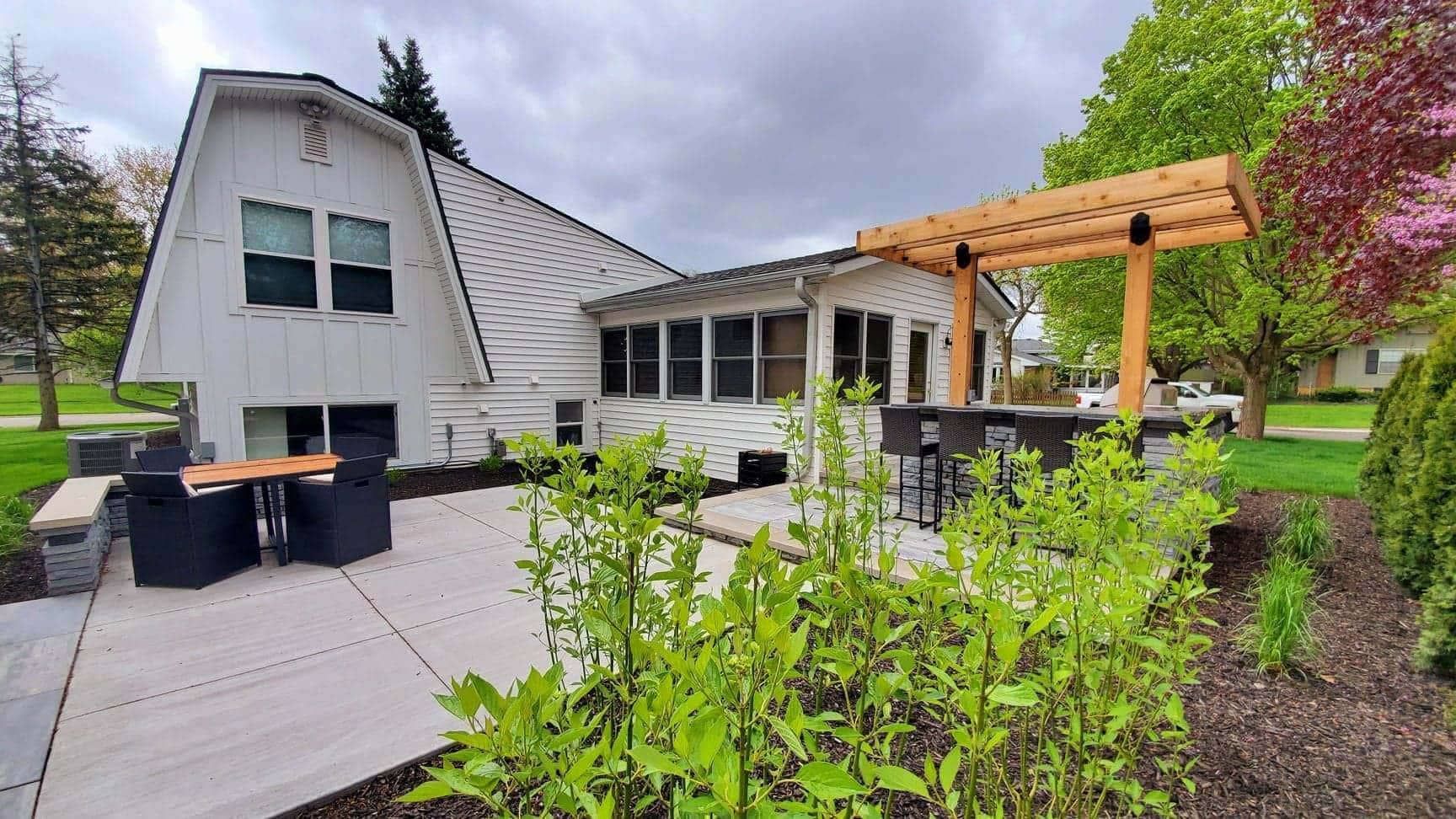 White house with outdoor patio, seating, and a wooden pergola. Green bushes in foreground. Cloudy sky.
