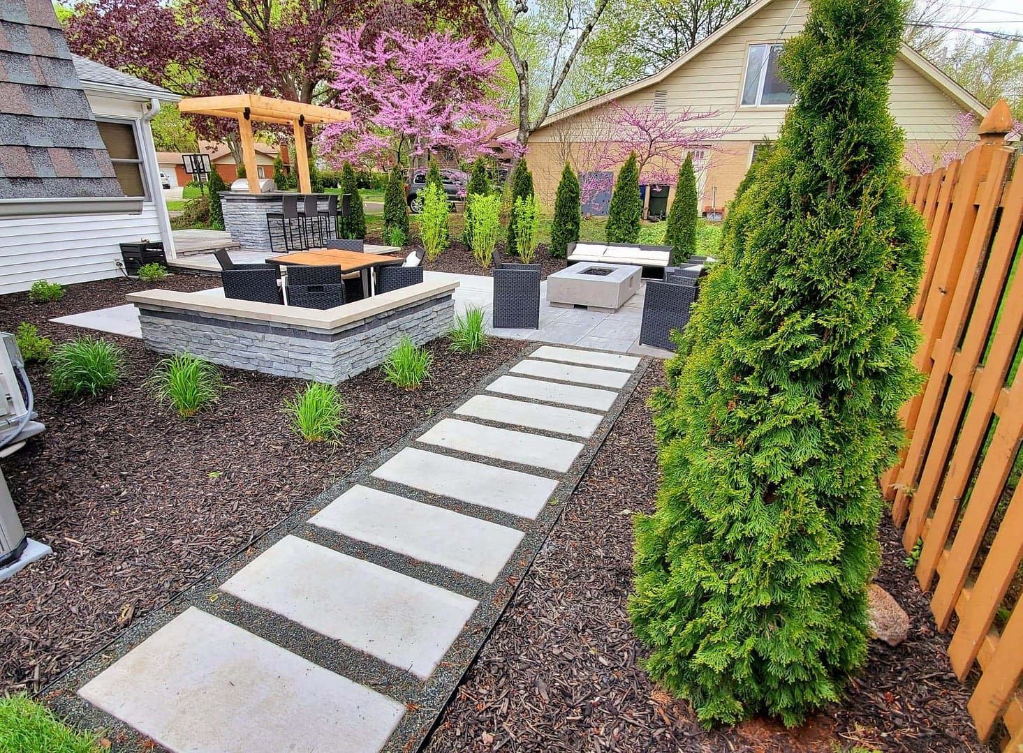 A modern outdoor patio with a stone pathway, fire pit, seating, and trees.