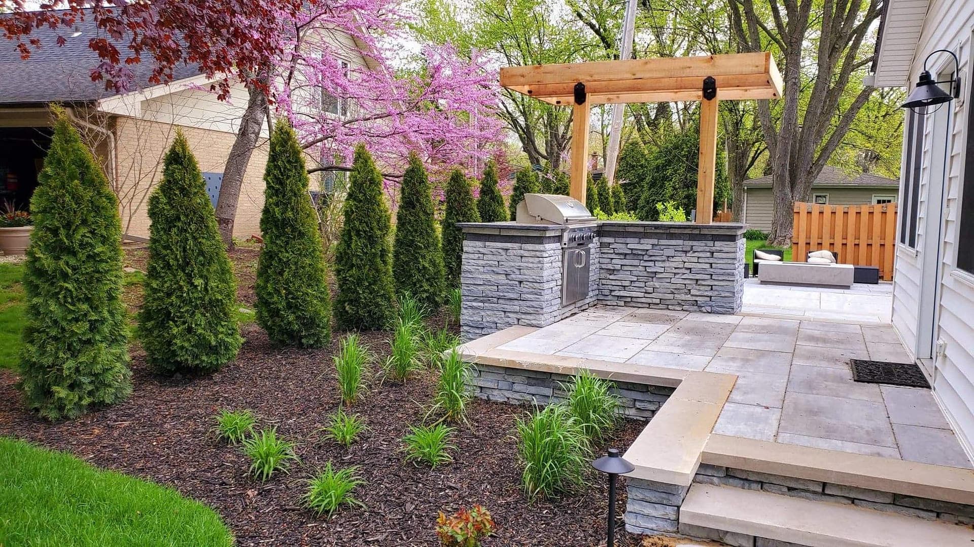 Landscaped backyard with a built-in grill, patio, and evergreen trees.