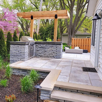 Backyard patio with built-in grill, stone and wood features, surrounded by greenery and a house.