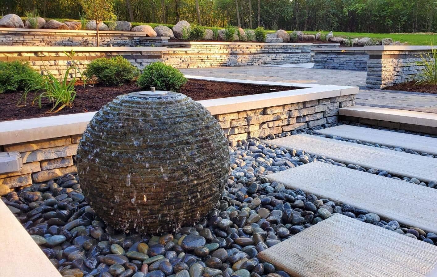 Stone sphere water fountain in a modern garden with stone walls, stepping stones, and river rock.