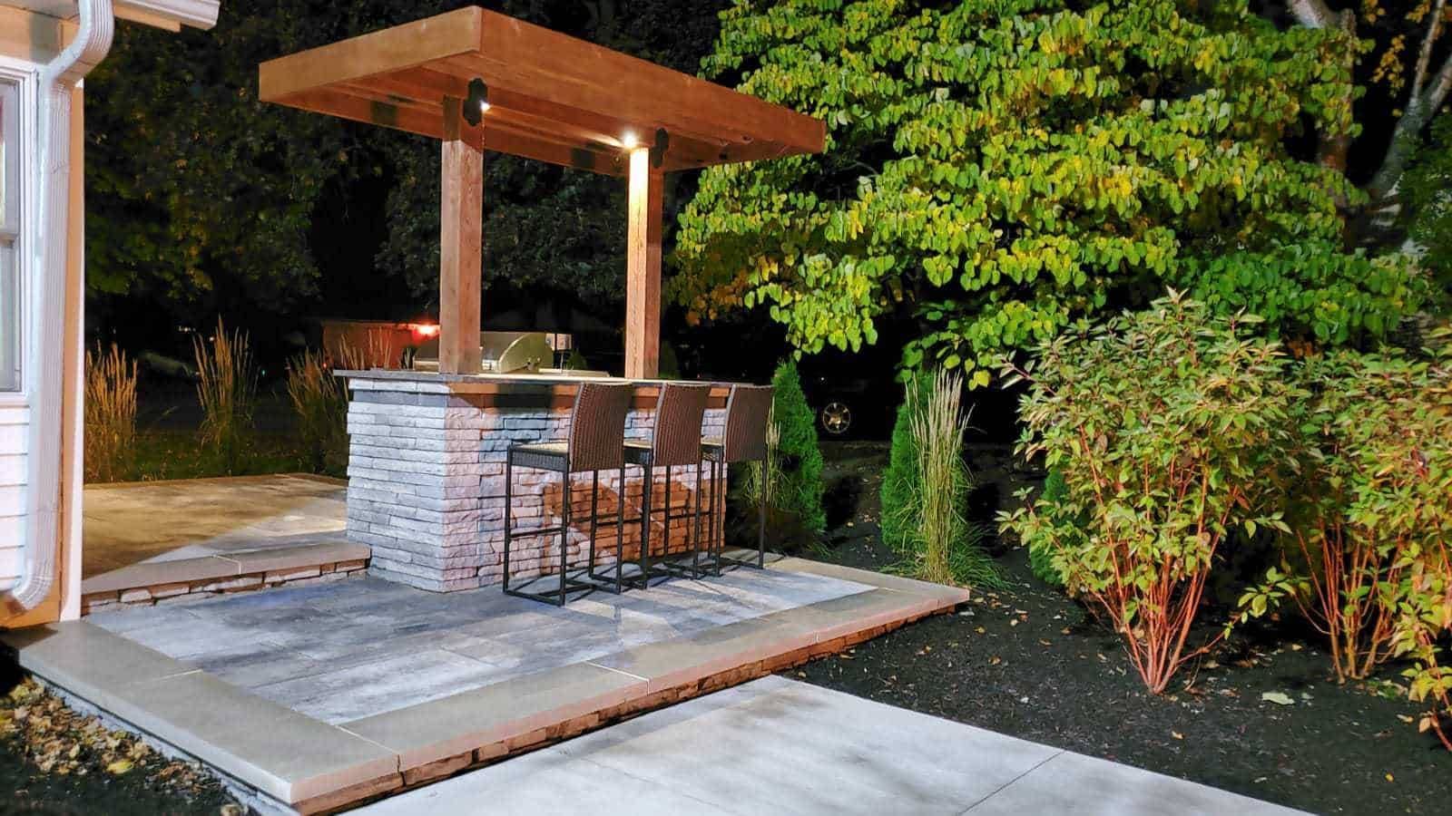 Outdoor bar with wooden canopy, brick facade, seating, and surrounding landscaping at night.