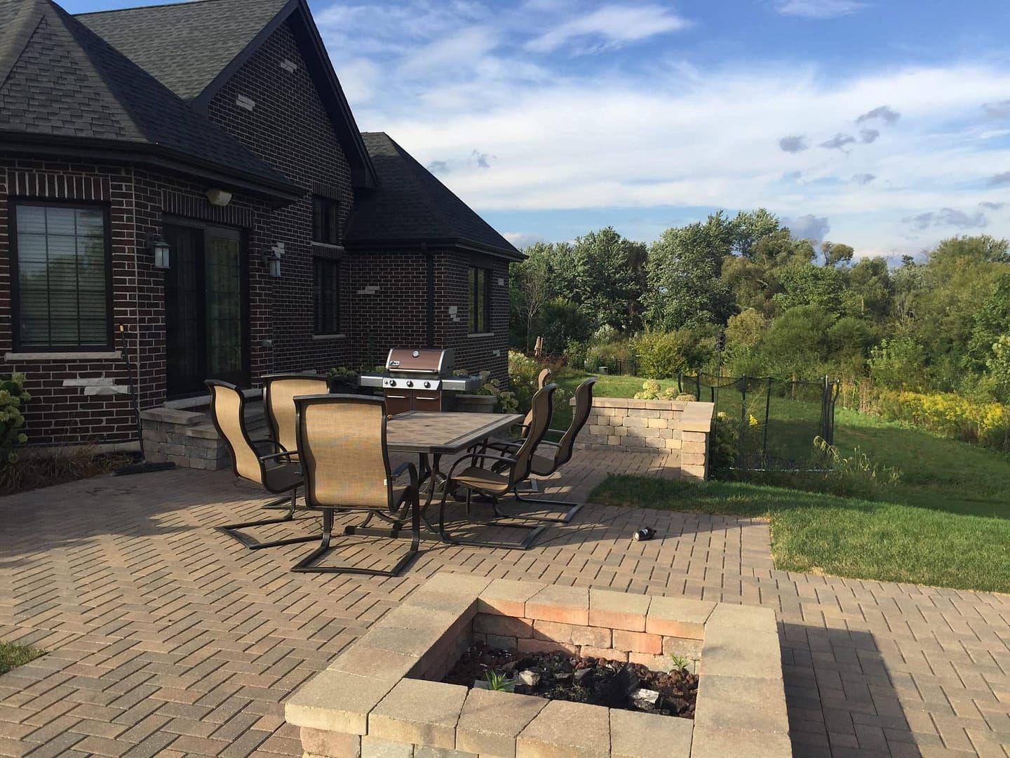 Brick patio with outdoor dining set, grill, and fire pit, overlooking a grassy area with trees.