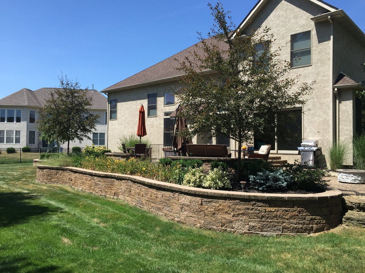 A stone retaining wall with landscaped flowerbeds and a patio adjoins a beige house on a sunny day.