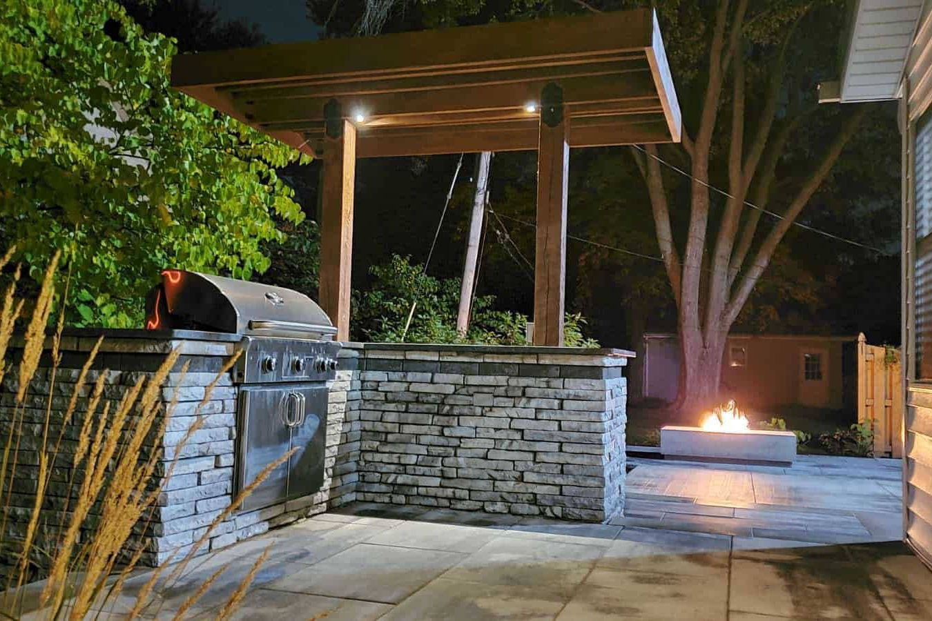 Outdoor kitchen with grill, stone counter, and fire pit at night. Pergola overhead.