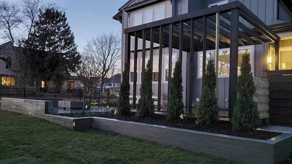 Modern home exterior with a dark pergola, tall green trees, and a long, low planter.
