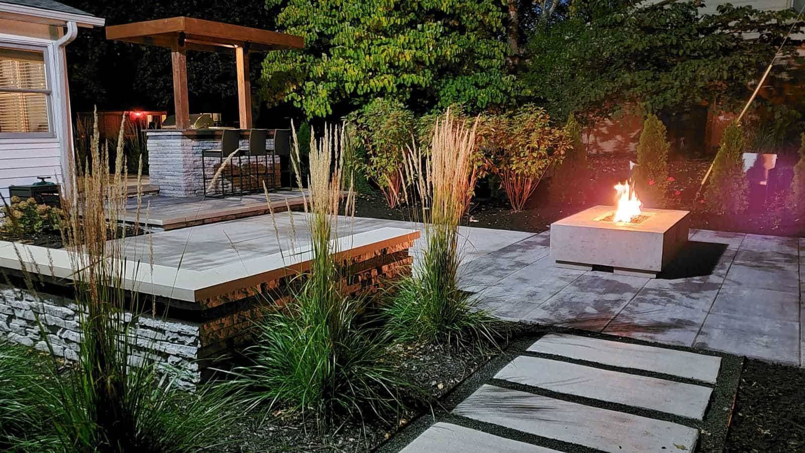 Outdoor patio with fire pit, stone pavers, and a bar area with a pergola, illuminated at night.