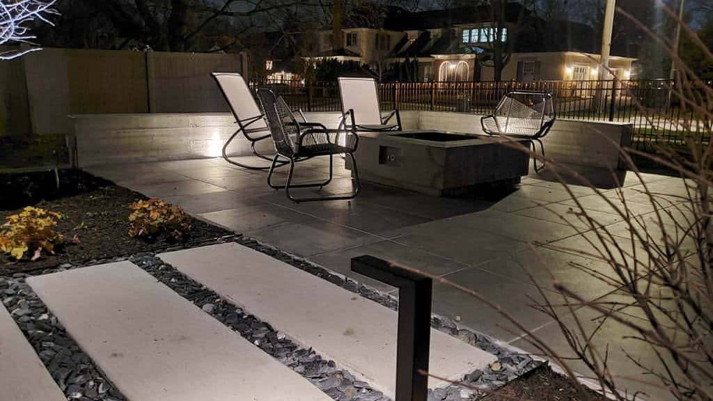 Night view of a modern patio with a fire pit, chairs, and stepping stones with landscape lighting.