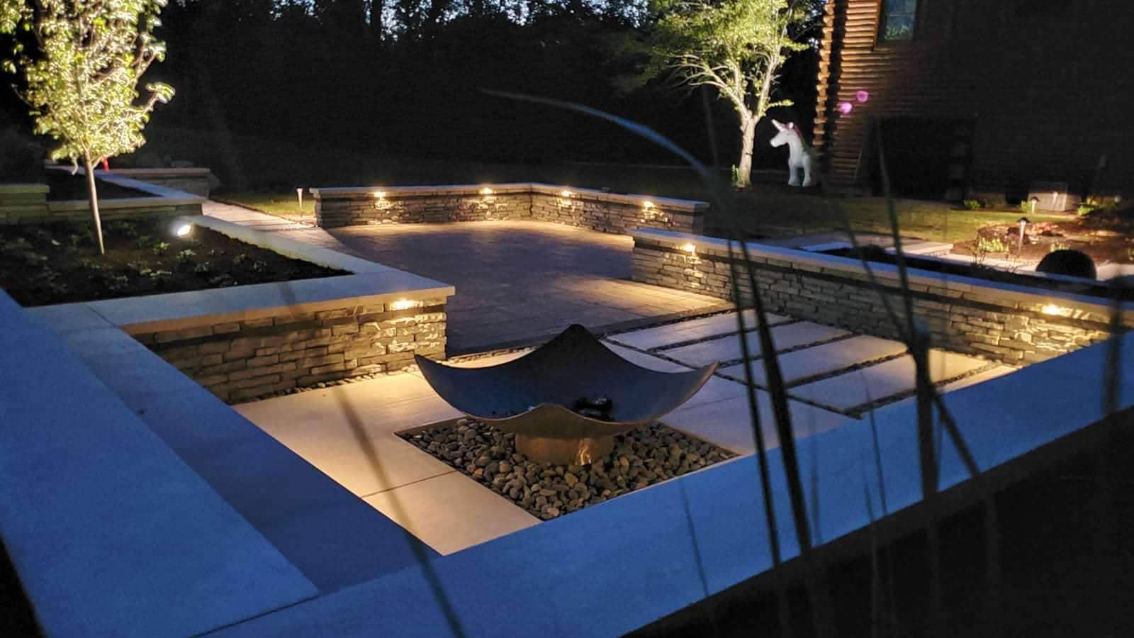 Nighttime outdoor patio with built-in lighting illuminating stone walls, square pavers, and a sculpture.