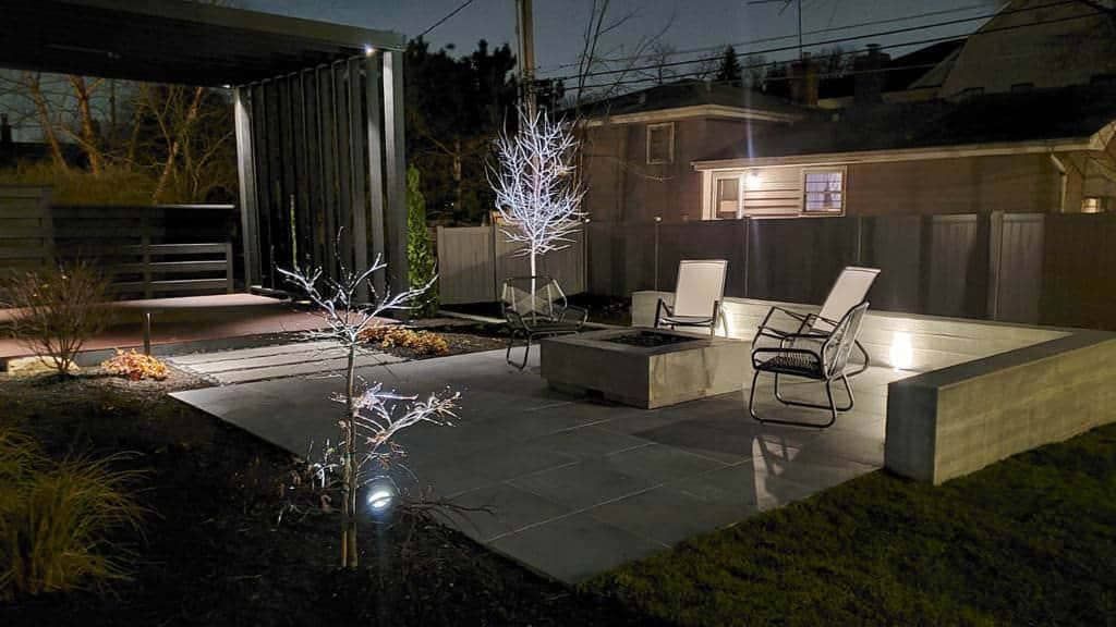 Nighttime backyard patio with fire pit, seating, and a modern pergola.