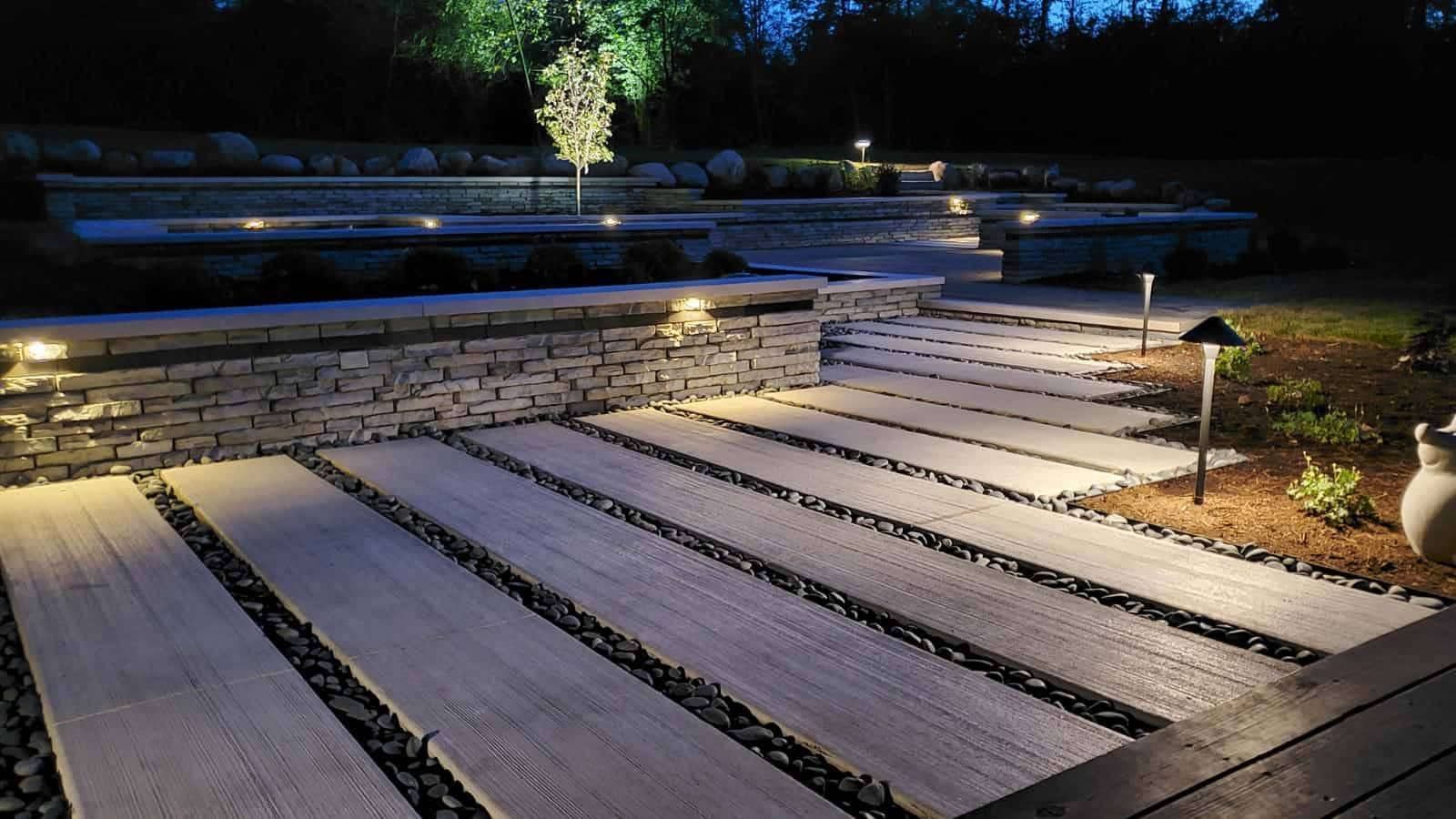 Lit stone pathway at night; rectangular stones with black gravel in between, leading to raised patio.