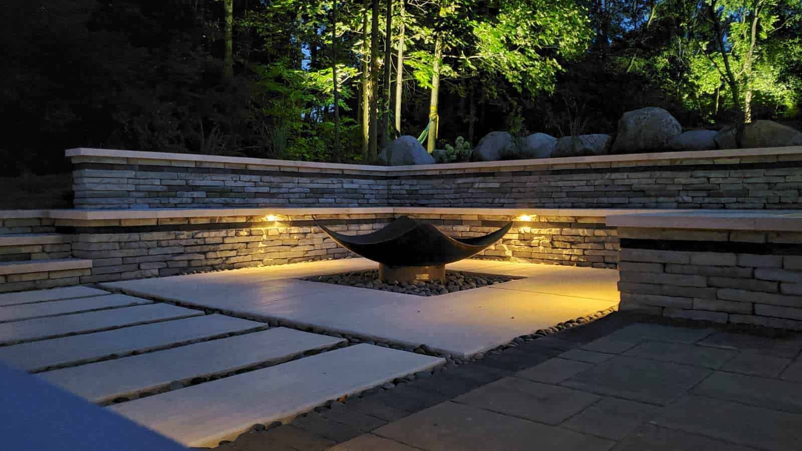 Stone patio with fire pit, lit by warm lights, surrounded by stone walls and trees at night.