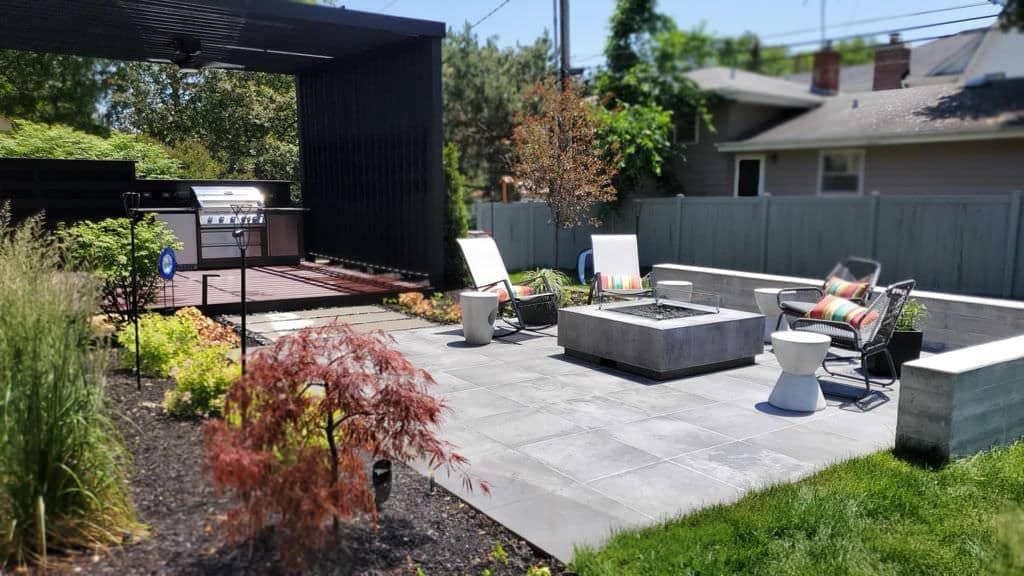 Backyard patio with grill, fire pit, seating, and landscaping on a sunny day.
