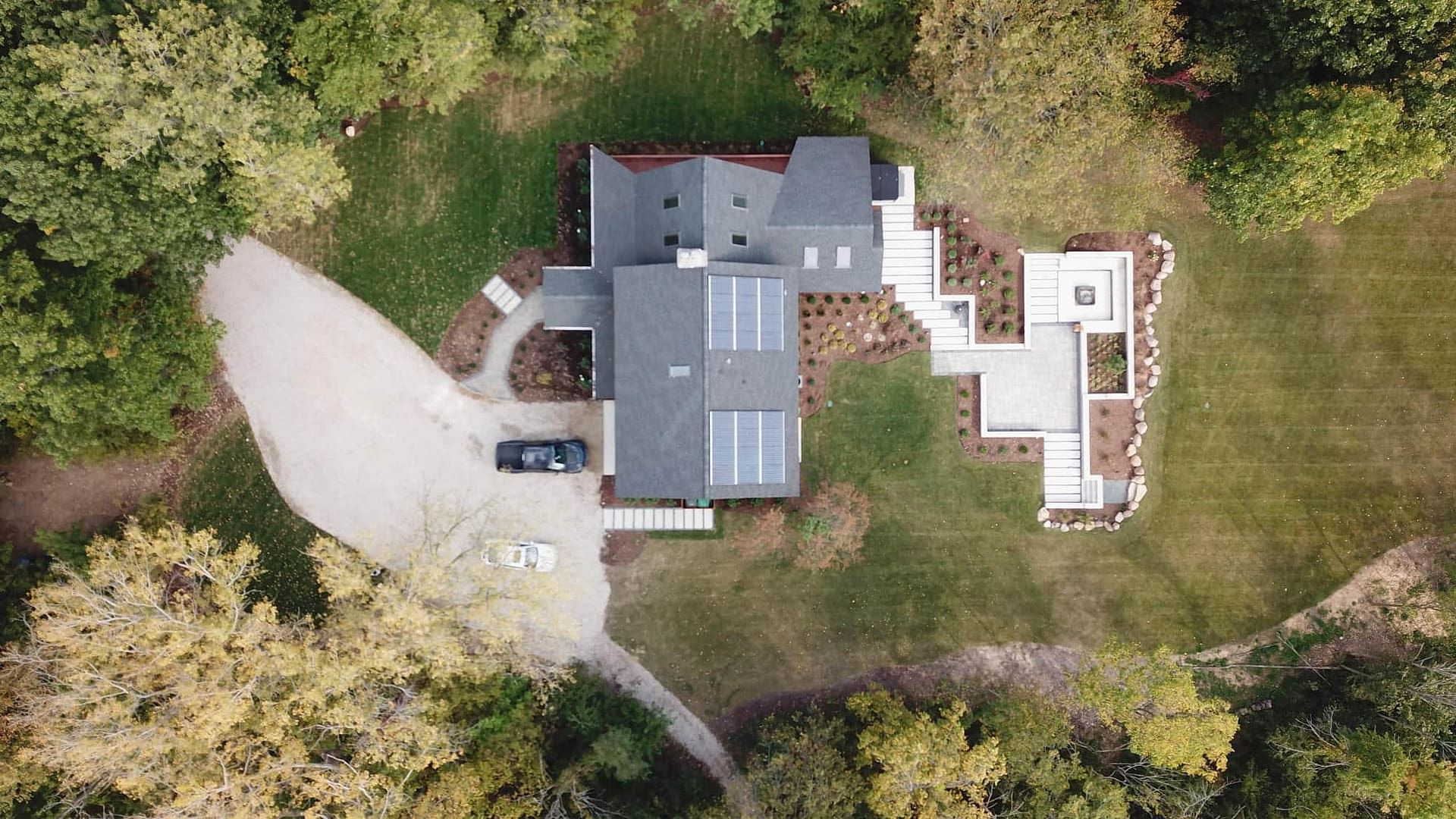 Aerial view of a modern house with solar panels, surrounded by trees and a gravel driveway.