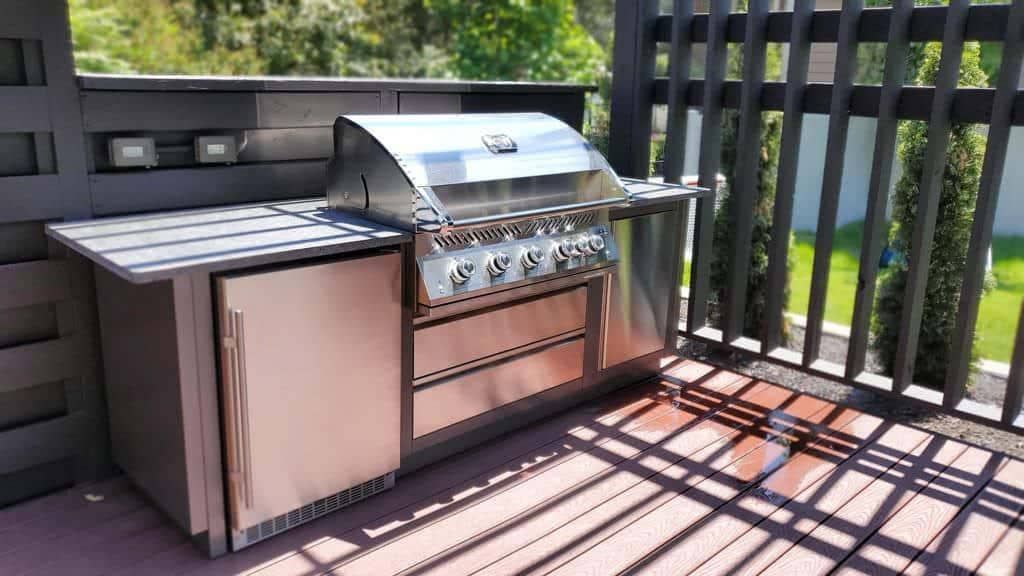 Stainless steel outdoor kitchen with grill, fridge, and workspace on a deck.