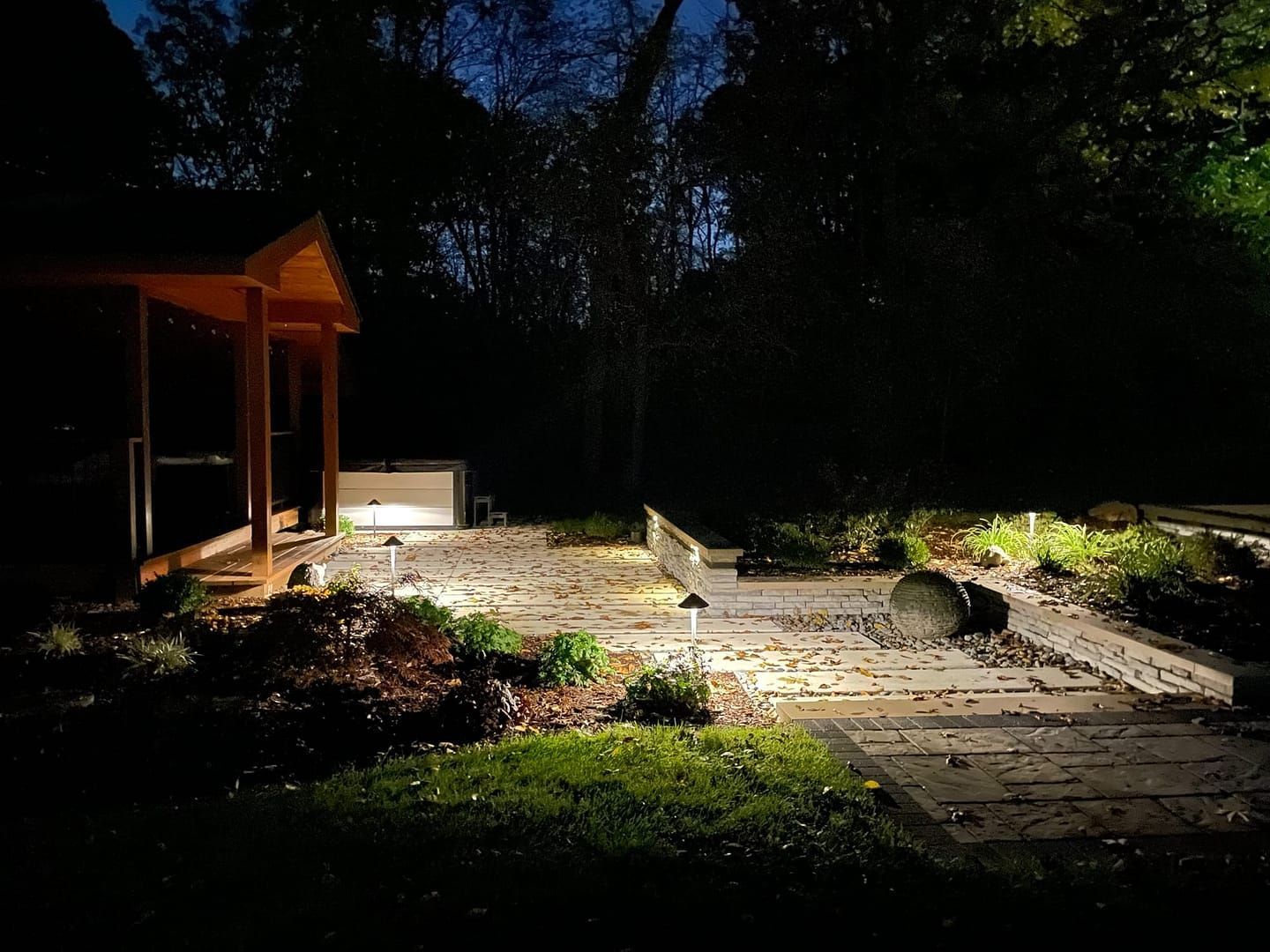 Night view of a lit patio with a wooden shelter, surrounded by trees and landscape lighting.