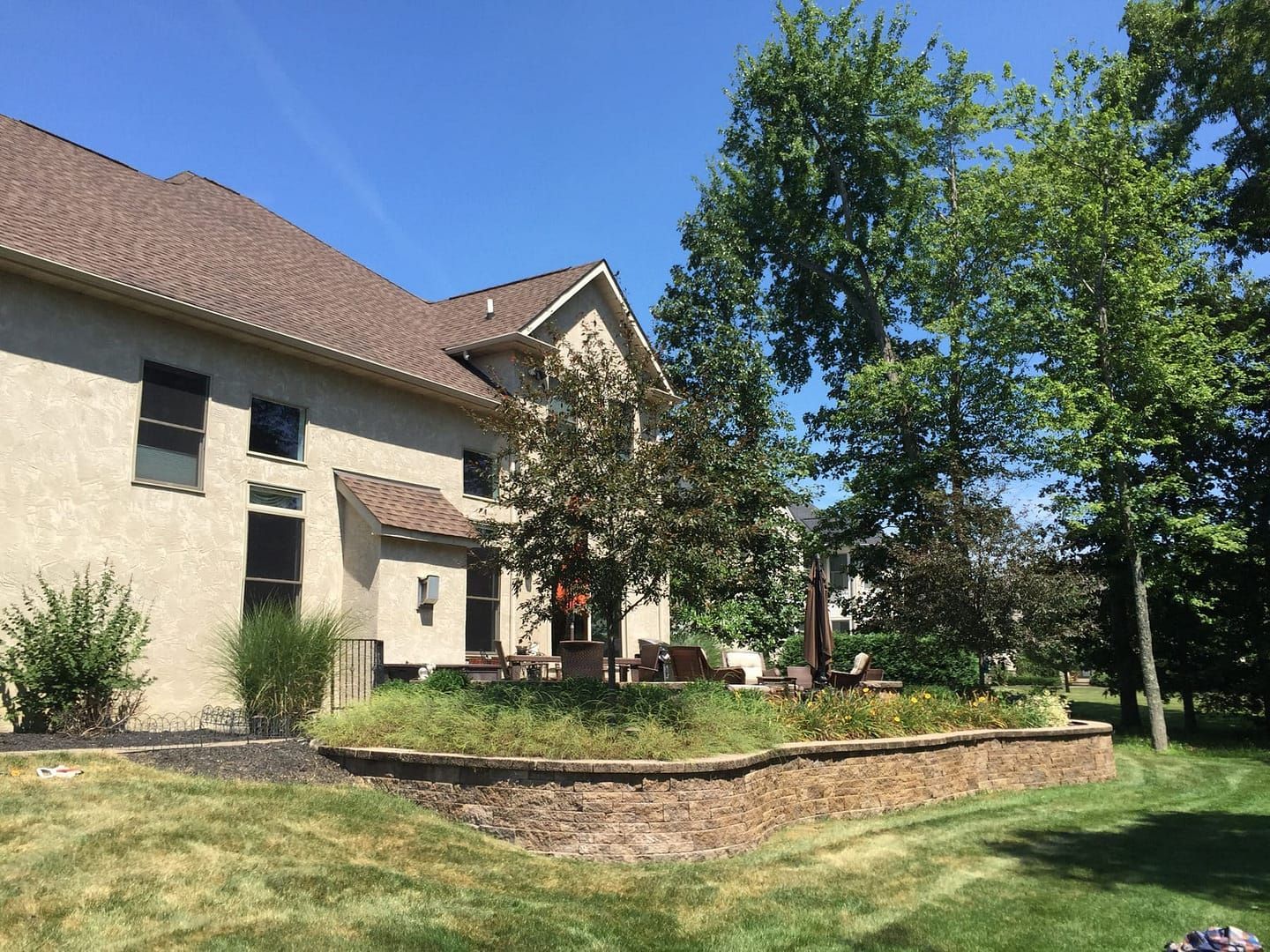 Backyard of a tan house with a stone retaining wall, trees, and a patio on a sunny day.