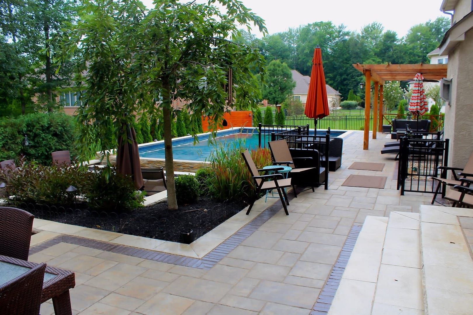 Patio with pool, chairs, umbrellas. Beige paving, greenery, and a wooden pergola.