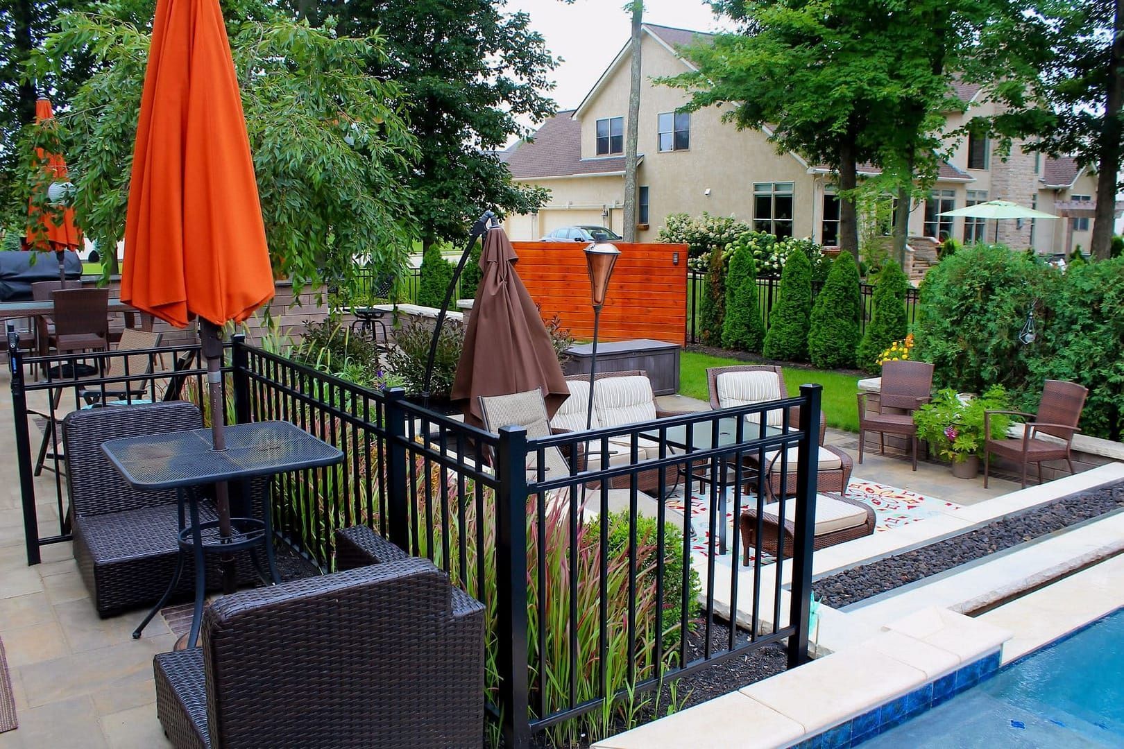 Outdoor patio with seating, umbrellas, and a pool. Brown and orange hues, with green trees and a house in the background.