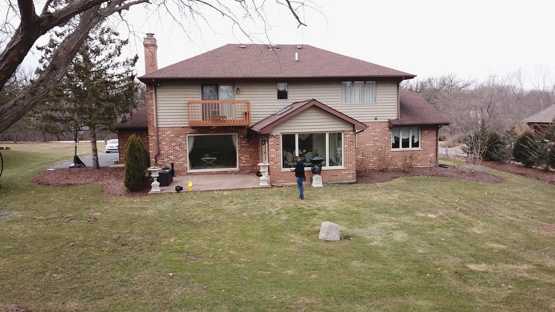 Two-story brick house with a person in the backyard; the yard has patchy grass and trees in the background.
