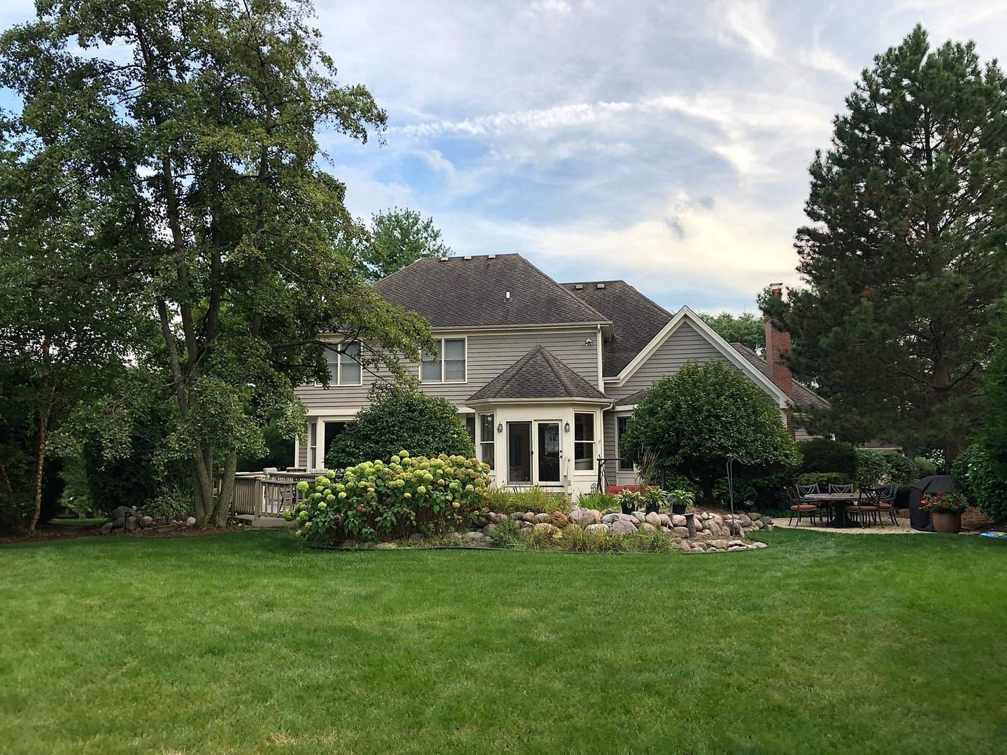 Large house with a light-colored exterior, surrounded by greenery, under a partly cloudy sky.