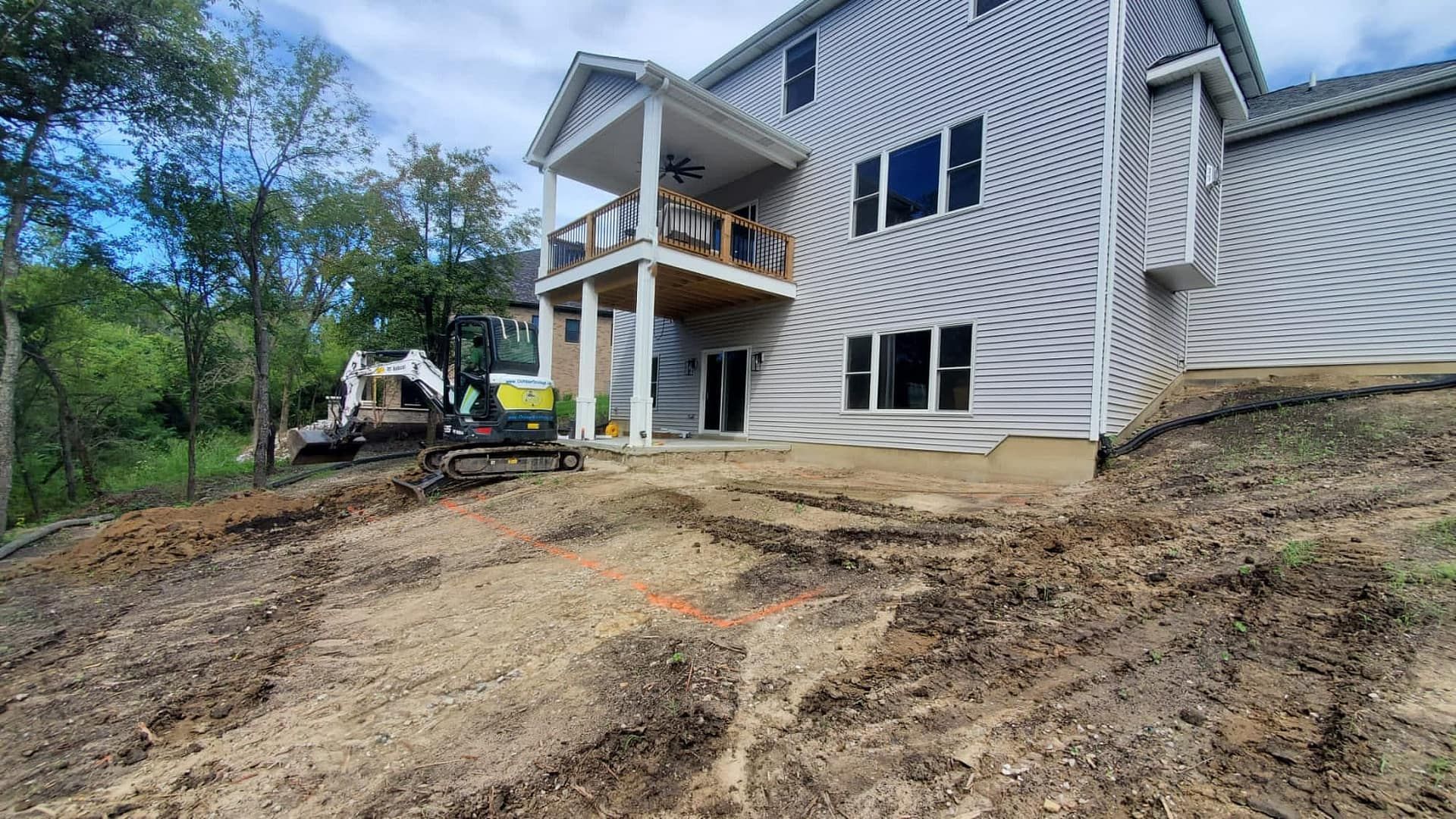 Backyard construction site; house with deck; muddy ground, excavator, trees in background.