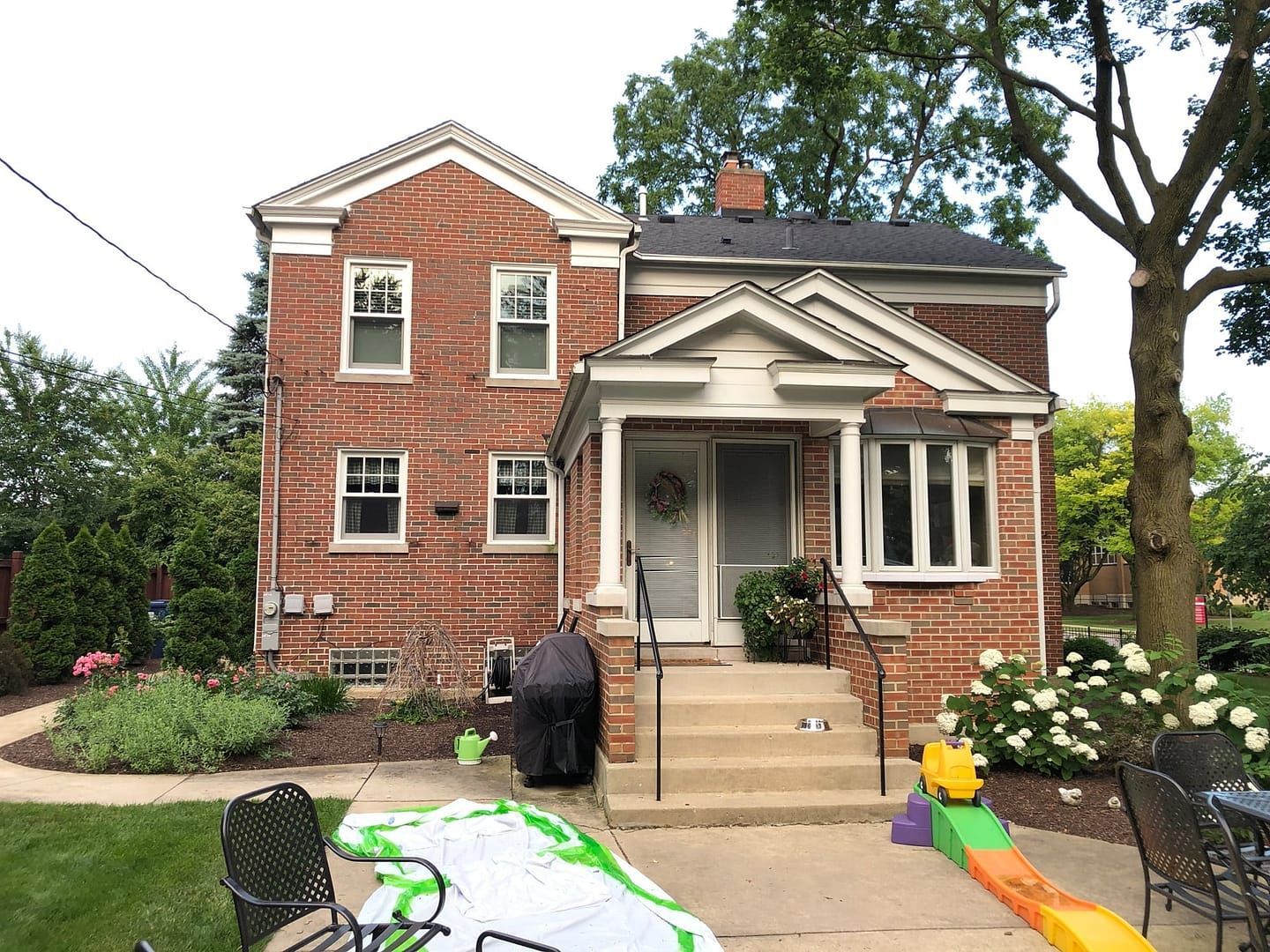 Two-story brick house with porch. Green lawn, flowers, and trees in yard.