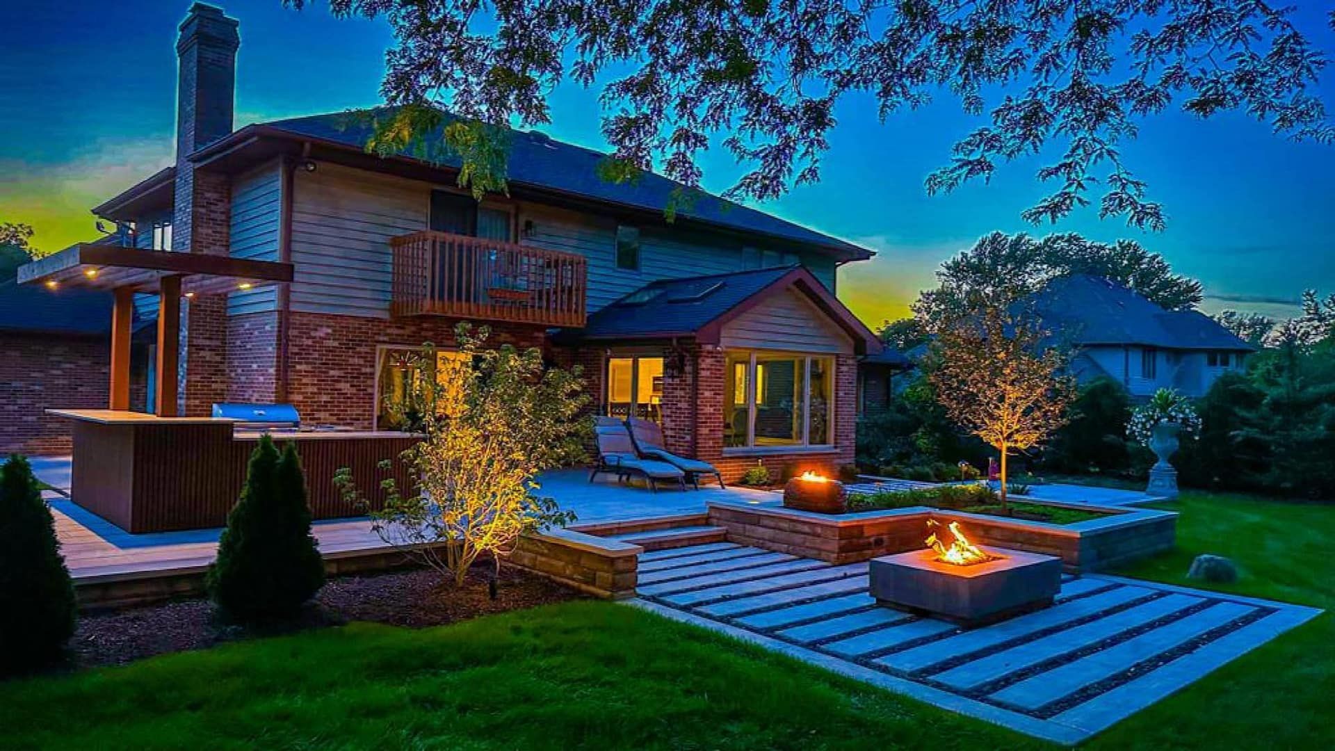 Backyard with a brick house at dusk, featuring a patio, fire pit, and outdoor bar, illuminated by warm lighting.