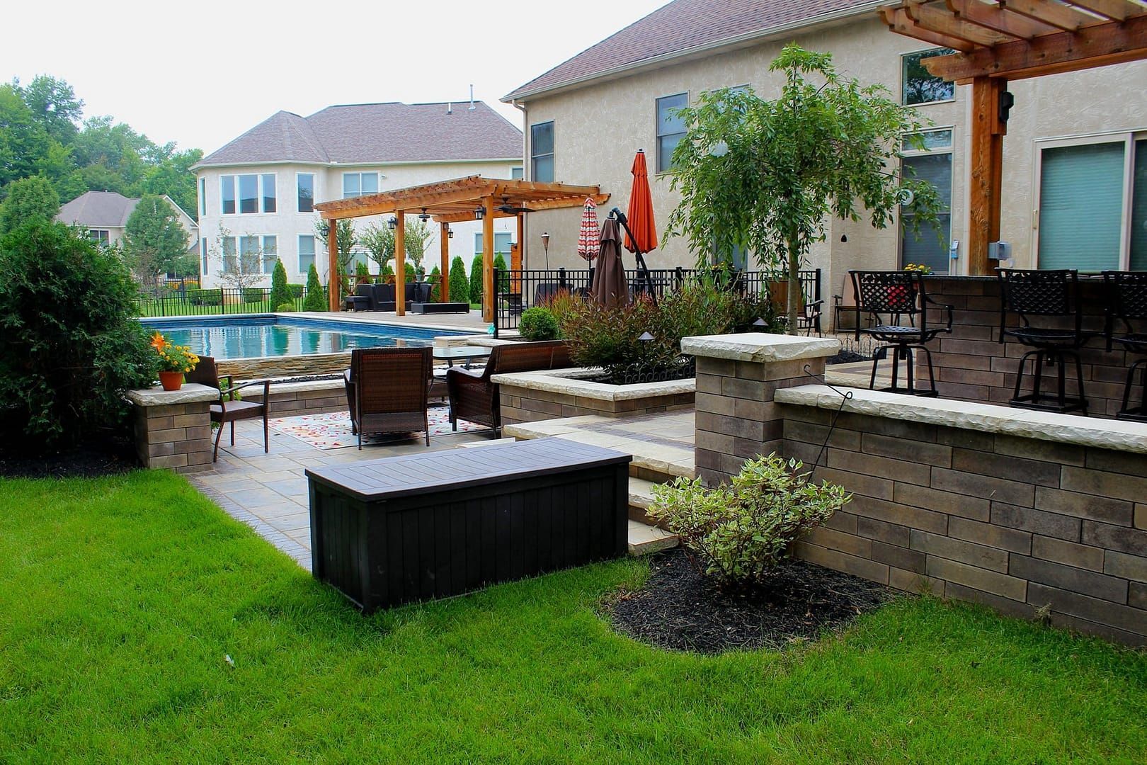 Backyard patio with pool, bar, and seating areas. Features a pergola, stonework, and green lawn.
