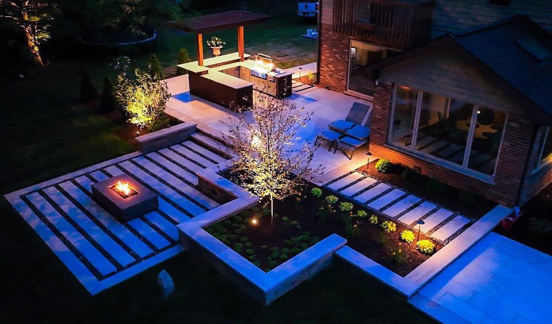 Night view of a landscaped backyard with a fire pit, patio, seating area, and garden beds, lit by outdoor lights.
