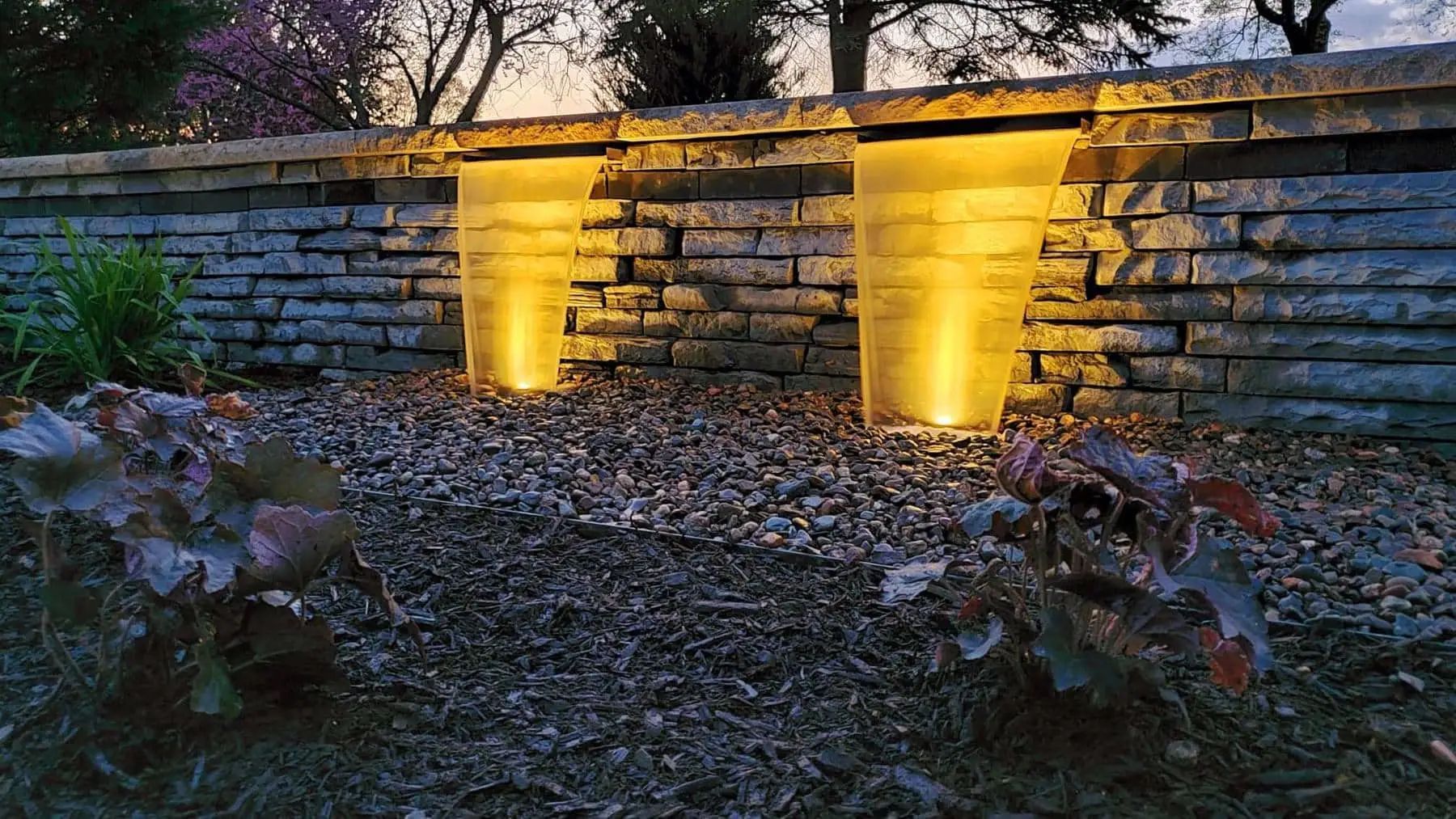 Two illuminated water features cascading from a stone wall. Dark, gravel ground and plants in foreground.