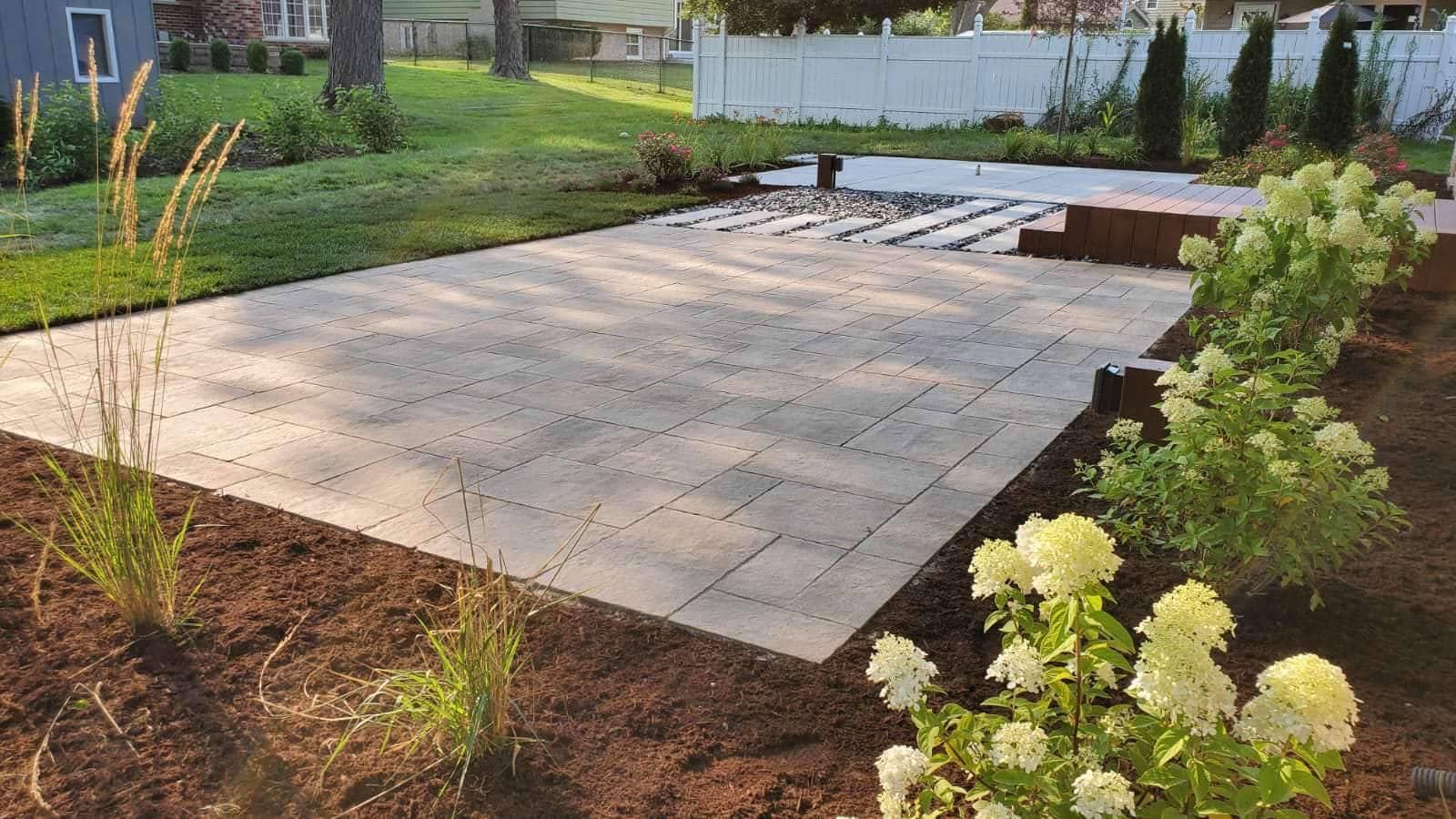 Large paved patio with landscaping, green lawn, and white fence.