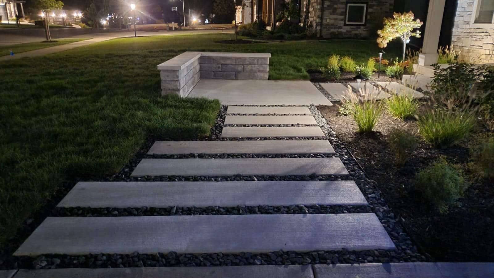 Nighttime view of a modern walkway with stepping stones, surrounded by grass and landscaping, lit by outdoor lights.