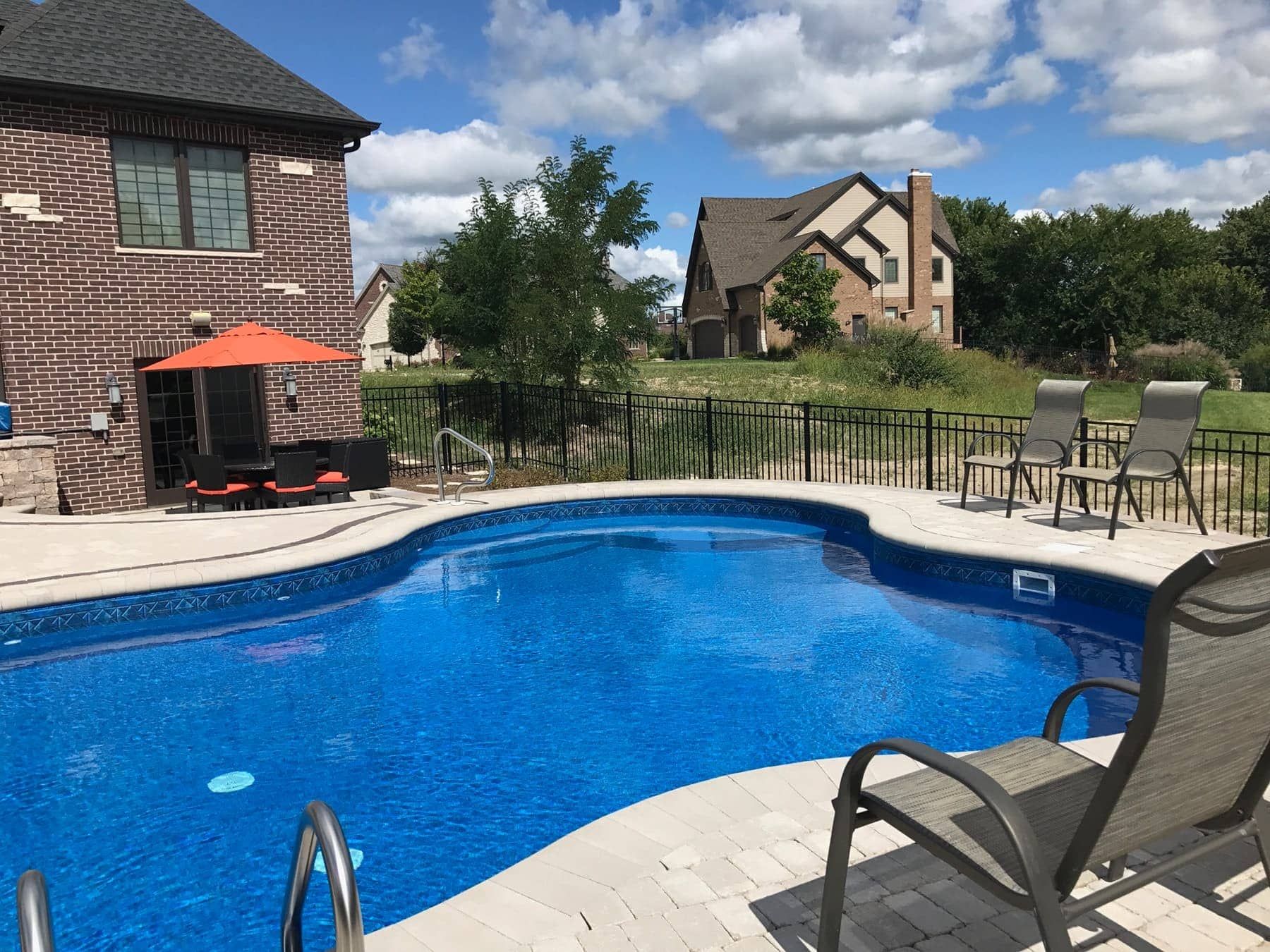 Swimming pool in a backyard with patio furniture and two-story brick house on a sunny day.