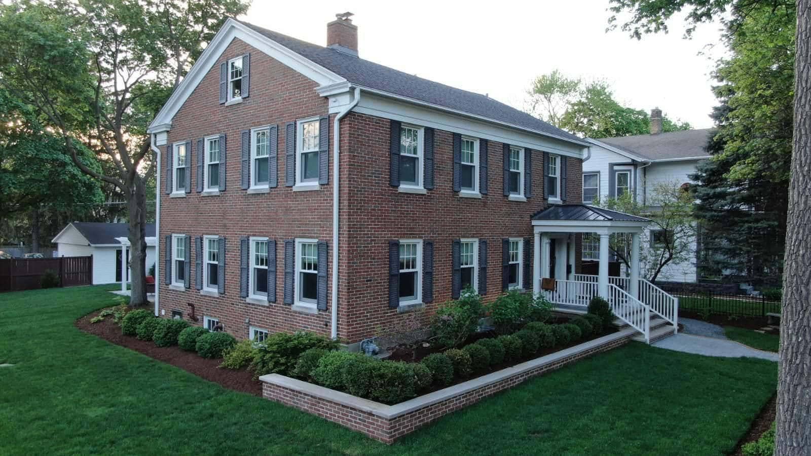 Two-story brick house with dark shutters, white trim, porch, and manicured lawn.