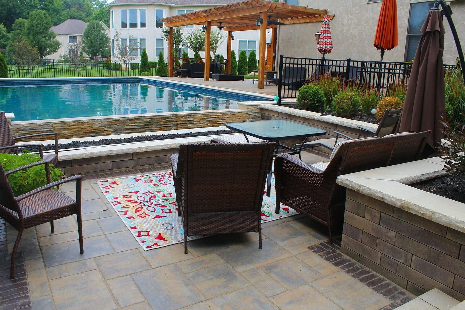 Patio with pool, pergola, outdoor furniture, rug, and umbrella in a backyard setting.