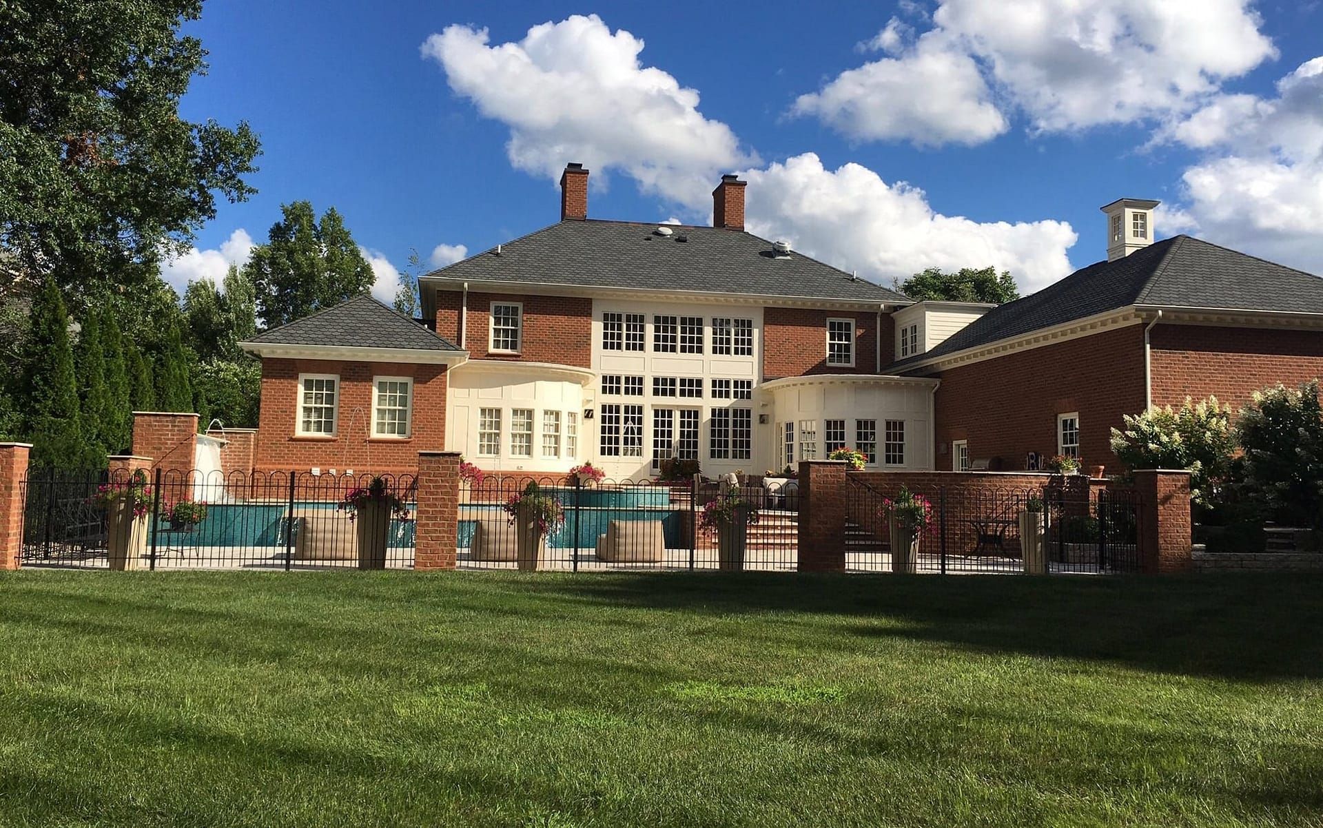 Large brick house with pool, green lawn, and blue sky with puffy white clouds.
